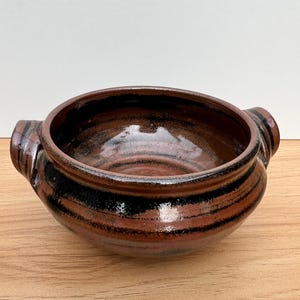 May include: A handmade ceramic bowl with a brown and black striped design. The bowl has two small handles and a rounded shape. The interior is a darker brown colour.