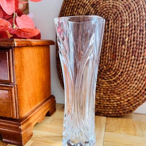 Könnte beinhalten: Eine klare Glasvase mit einem strukturierten, wirbelnden Design. Die Vase hat eine ausgestellte Form und steht auf einer Holzoberfläche. Der Hintergrund umfasst einen Holzschrank und einen gewebten kreisförmigen Wandbehang.