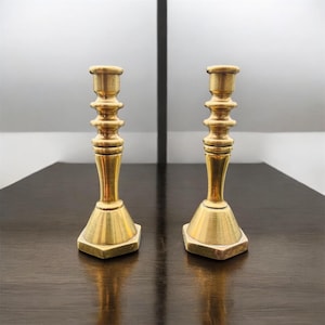 Könnte beinhalten: Zwei Messing-Kerzenhalter mit sechseckigem Sockel und abgestuftem Design. Die Kerzenhalter haben einen warmen, goldenen Farbton und werden auf einer dunklen Holzoberfläche präsentiert. Das Design ist klassisch und elegant.
