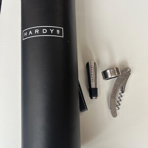 May include: A black wine bottle carrier with the word "HARDYS" in white. Includes a wine stopper, a bottle ring, and a corkscrew. The wine accessories are silver-coloured.