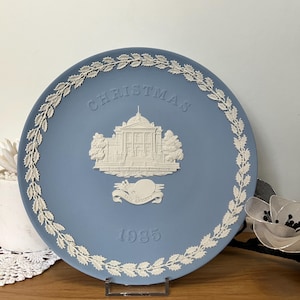 Puede incluir: Un plato decorativo azul con un diseño en relieve blanco. El plato presenta un edificio, la palabra "CHRISTMAS" y el año "1985". Un borde de guirnalda blanca rodea el borde del plato. El plato se exhibe en un soporte de metal.