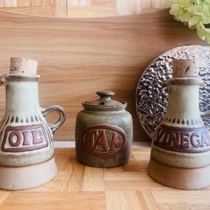 Puede incluir: Un conjunto de tres recipientes de cocina de cerámica. Dos botellas con tapones de corcho y asas están etiquetadas con "OIL" y "VINEGAR" en letras marrones. Un pequeño tarro con tapa está etiquetado con "JAM". Los recipientes son beige y marrón.