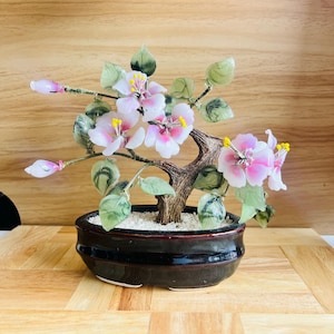 以下が含まれることがあります： ピンクと白の花、黄色の雄しべ、緑の葉を持つ装飾的な盆栽。木は濃い茶色のセラミックポットに入っており、木の背景に配置されています。盆栽はユニークな室内装飾品です。