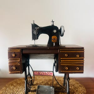 May include: A miniature black sewing machine with a silver wheel, mounted on a brown wooden cabinet featuring gold-toned drawer accents and a central metal treadle. A small rectangular sign with the word SEWING is attached to the front frame.