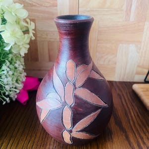 May include: A dark brown ceramic vase with a narrow neck and a rounded body, featuring a carved bamboo leaf pattern in a lighter terracotta colour. The surface has a textured, matte finish with visible brush strokes and a dark rim at the opening.
