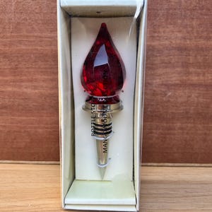 May include: A red, teardrop-shaped bottle stopper with a silver base, presented in a white box. The stopper has the text "Bottle Stopper" and "Mango Moon" printed on it. The stopper is designed to seal wine bottles.