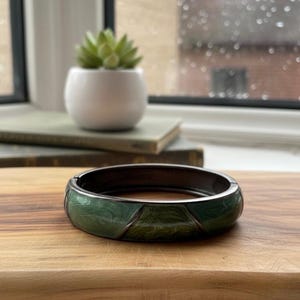 Op de afbeelding: Een donker metalen scharnierende armband met een groen emaille ontwerp. De armband heeft een geometrisch patroon met groentinten en wordt gepresenteerd op een houten snijplank. Op de achtergrond staat een kleine vetplant in een witte pot.