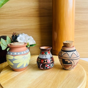 May include: Three small terracotta vessels with distinct hand-painted designs. The left pot features yellow petals and white dots. The center vessel has a geometric red, black, and white pattern. The right pot has a cork stopper and brown geometric motifs.