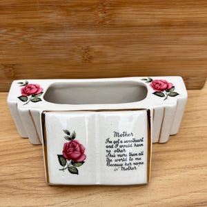Pode incluir: Um vaso de cerâmica branco com formato retangular e acabamento dourado. O vaso apresenta ilustrações de rosas cor-de-rosa e o texto "Mother" com um poema. É um item decorativo.