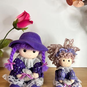 May include: Two handmade fabric dolls. The larger doll wears a purple hat, dress with white lace trim, and has purple braided hair. The smaller doll has curly hair and a similar dress. Both dolls have smiling faces and are on a wooden surface.