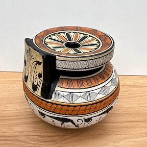 Puede incluir: Un jarrón de cerámica pintado a mano con tapa. El jarrón está decorado con patrones geométricos y figuras en negro, marrón y blanco. La tapa presenta un diseño floral. El jarrón mide unos 13 cm de alto.