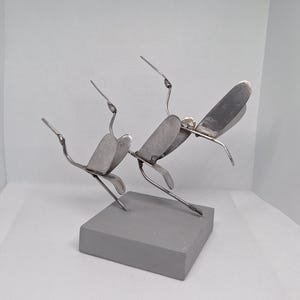 May include: A metal sculpture depicting three birds in flight, made from silver-coloured metal. The birds are mounted on a rectangular, grey concrete base. The wings are made from flat, curved metal pieces.