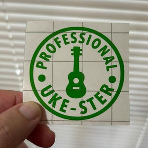May include: A square sticker with a green circular design featuring a ukulele silhouette and the words "PROFESSIONAL UKE-STER" in green. The sticker is on a white background with a grid pattern.