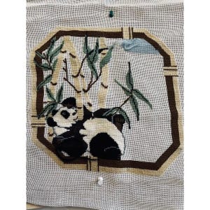 May include: Needlepoint of a panda bear resting among bamboo stalks. The panda is black and white, set within a brown octagonal frame. Green leaves and a light blue cloud accent the scene. The background is a light beige fabric.