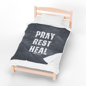 Puede incluir: Una cama con estructura de madera clara y ropa de cama blanca. Una manta gris oscuro con las palabras "PRAY REST HEAL" en letras blancas está sobre la cama. El texto "PSALM 23:2-3" está debajo.