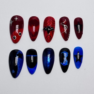 Pode incluir: Um conjunto de dez unhas postiças em formato de amêndoa. A fileira superior apresenta unhas vermelhas com detalhes de estrelas prateadas, um pingente de morcego preto e linhas prateadas. A fileira inferior apresenta unhas azuis e pretas.