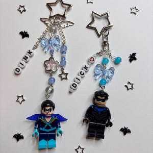 Puede incluir: Dos llaveros hechos a mano con figuras de superhéroes. Cada llavero tiene una parte superior plateada en forma de estrella con alas de ángel, cuentas azules, un lazo y una pequeña figura de superhéroe. Un llavero deletrea "DINK" y el otro "DICK".