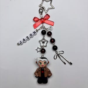May include: A handmade keychain with a small figurine of a character in a brown jacket and black trousers. The keychain includes a red bow, star-shaped charms, and beads. The name "JASON" is spelled out in white letter beads.