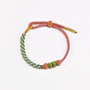 Puede incluir: Una pulsera hecha a mano con un diseño trenzado rosa y verde. La pulsera tiene un cierre de nudo corredizo y detalles dorados. La sección trenzada es verde y rosa, con una serie de nudos verdes y rosas.