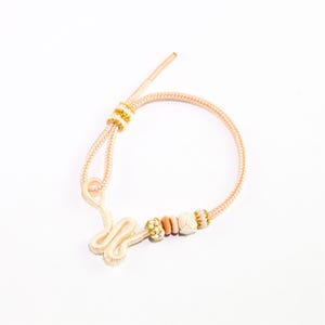 May include: A pink and white braided bracelet with a knot design and beads. The bracelet has a white and gold striped accent.