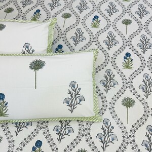 Puede incluir: Juego de cama blanco con un estampado floral en azul y verde. El juego incluye fundas de almohada con un borde a rayas verdes y diseños florales a juego. El fondo presenta un patrón repetitivo de flores y hojas estilizadas.