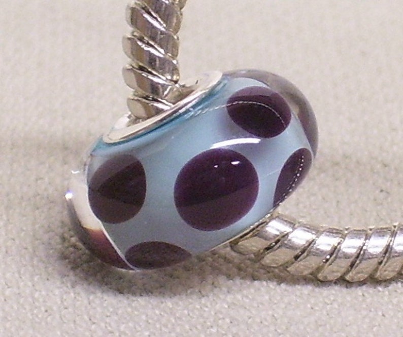 Pode incluir: Um bracelete de prata com um berloque de vidro azul claro com bolinhas roxas.