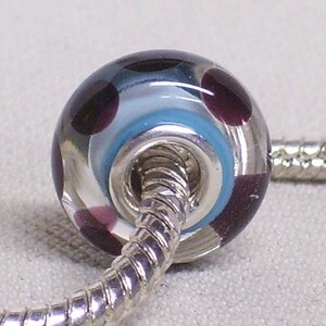 Pode incluir: Uma conta de vidro transparente com um n&uacute;cleo de metal prateado. A conta &eacute; decorada com redemoinhos de azul, branco e roxo.