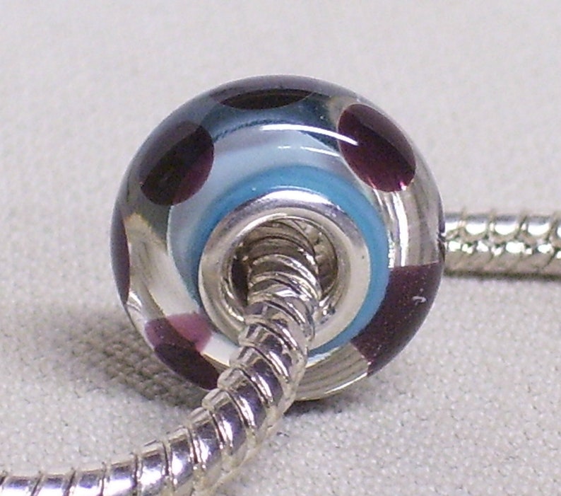 Pode incluir: Uma conta de vidro transparente com um n&uacute;cleo de metal prateado. A conta &eacute; decorada com redemoinhos de azul, branco e roxo.