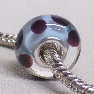 Pode incluir: Uma conta de vidro azul claro com bolinhas roxas escuras. A conta tem um n&uacute;cleo de metal prateado e &eacute; enfiada em uma corrente prateada.