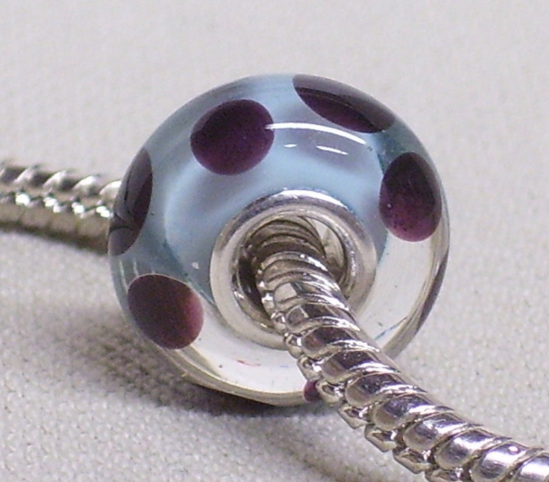 Pode incluir: Uma conta de vidro azul claro com bolinhas roxas escuras. A conta tem um n&uacute;cleo de metal prateado e &eacute; enfiada em uma corrente prateada.