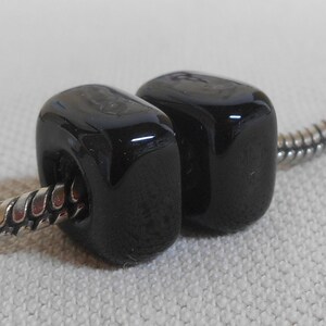 May include: Two glossy black beads with rounded edges are strung on a silver chain. The beads have a smooth, reflective surface, and are close together. The background is a light, textured fabric.