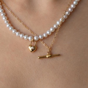 May include: A layered necklace featuring a strand of small white pearls and a delicate gold-tone chain. The necklace includes a gold-tone heart-shaped padlock charm with a tiny pearl inlay and a gold-tone toggle clasp closure hanging at the center.