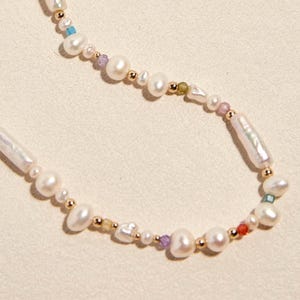 Op de afbeelding: Een ketting met een streng witte zoetwaterparels in diverse vormen, waaronder langwerpige en ronde exemplaren, afgewisseld met kleine goudkleurige metalen kralen en piepkleine gekleurde accentkralen in blauw, paars, groen en rood.