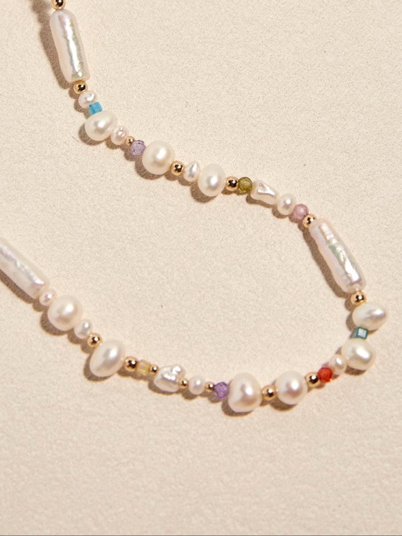 Op de afbeelding: Een ketting met een streng witte zoetwaterparels in diverse vormen, waaronder langwerpige en ronde exemplaren, afgewisseld met kleine goudkleurige metalen kralen en piepkleine gekleurde accentkralen in blauw, paars, groen en rood.