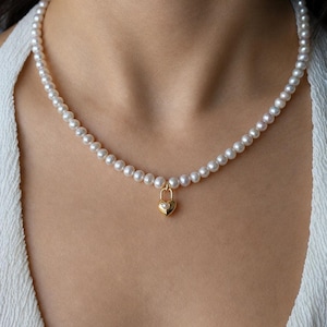 May include: A necklace featuring a strand of small, round, white pearls. A gold-toned heart-shaped padlock charm is suspended from the centre of the pearl strand. The padlock charm features a small, embedded white pearl at its centre.