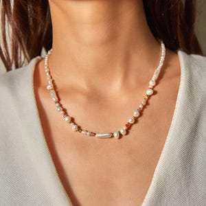 Op de afbeelding: Een ketting bestaande uit een streng witte zoetwaterparels in vari&euml;rende vormen en maten, afgewisseld met kleine, veelkleurige glaskralen en piepkleine goudkleurige metalen tussenstukjes, wat een delicaat en gestructureerd sieraad vormt.