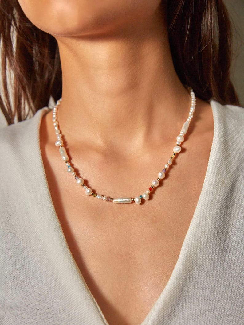 Op de afbeelding: Een ketting bestaande uit een streng witte zoetwaterparels in vari&euml;rende vormen en maten, afgewisseld met kleine, veelkleurige glaskralen en piepkleine goudkleurige metalen tussenstukjes, wat een delicaat en gestructureerd sieraad vormt.