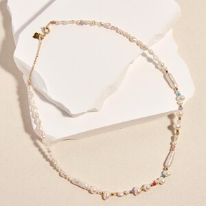 Op de afbeelding: Een ketting bestaande uit een streng onregelmatige witte zoetwaterparels, afgewisseld met kleine goudkleurige metalen kralen en piepkleine veelkleurige edelsteenaccenten. Het sieraad heeft een goudkleurige metalen sluiting en een klein hartvormig bedeltje.