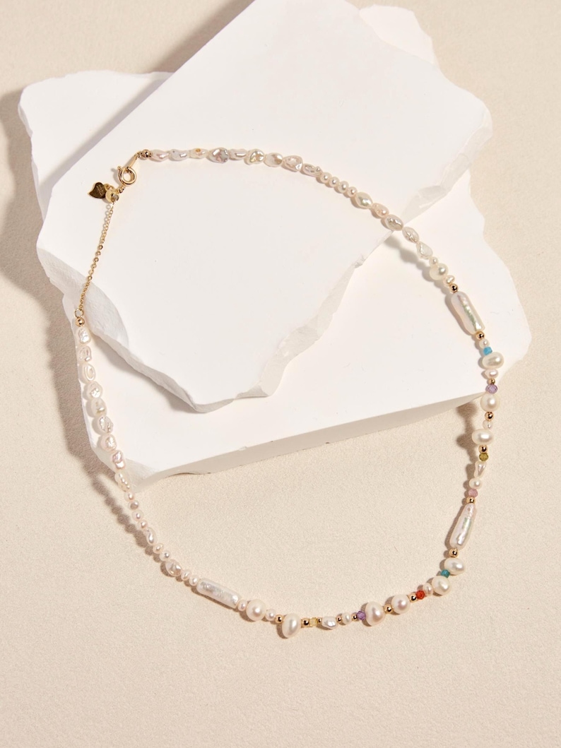 Op de afbeelding: Een ketting bestaande uit een streng onregelmatige witte zoetwaterparels, afgewisseld met kleine goudkleurige metalen kralen en piepkleine veelkleurige edelsteenaccenten. Het sieraad heeft een goudkleurige metalen sluiting en een klein hartvormig bedeltje.