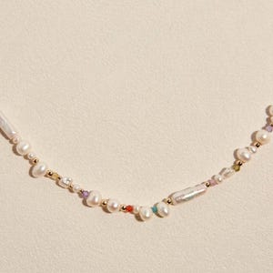 Op de afbeelding: Een ketting bestaande uit een streng witte zoetwaterparels in diverse vormen en maten, afgewisseld met kleine goudkleurige metalen kralen en piepkleine, kleurrijke gefacetteerde edelsteenaccenten in een asymmetrisch patroon.