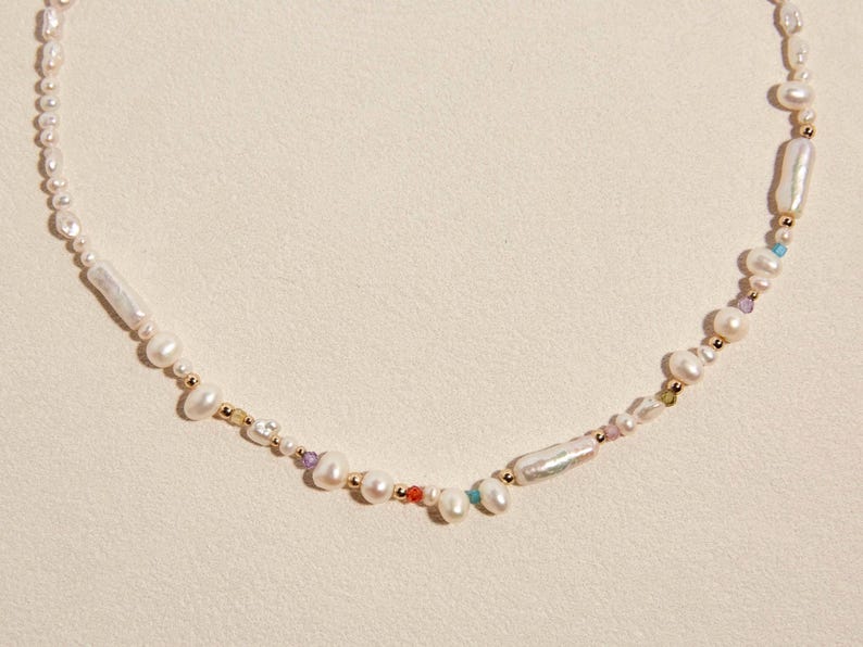 Op de afbeelding: Een ketting bestaande uit een streng witte zoetwaterparels in diverse vormen en maten, afgewisseld met kleine goudkleurige metalen kralen en piepkleine, kleurrijke gefacetteerde edelsteenaccenten in een asymmetrisch patroon.