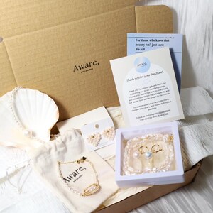 Op de afbeelding: Een kartonnen geschenkdoos met een parelketting gedrapeerd over een witte schelp, een gouden kettingarmband met parels op een beige stoffen zakje, hartvormige pareloorbellen op een kaart en een parelketting in een wit doosje met een briefje.