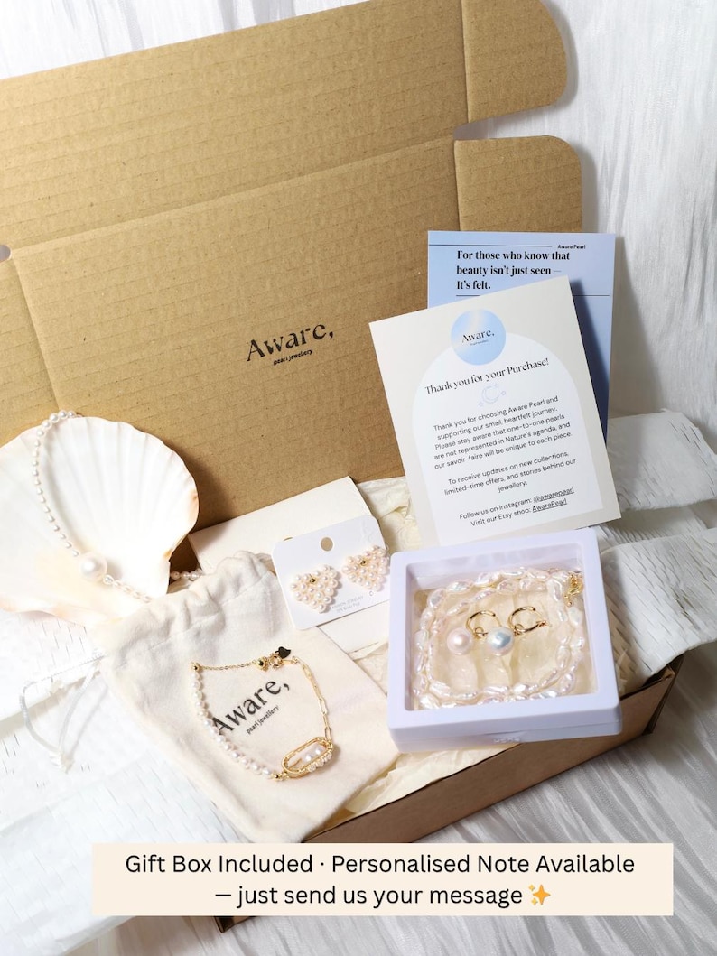 Op de afbeelding: Een kartonnen geschenkdoos met een parelketting gedrapeerd over een witte schelp, een gouden kettingarmband met parels op een beige stoffen zakje, hartvormige pareloorbellen op een kaart en een parelketting in een wit doosje met een briefje.