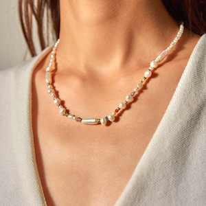 Op de afbeelding: Een ketting bestaande uit een streng witte zoetwaterparels afgewisseld met kleine, veelkleurige edelsteenkralen en piepkleine goudkleurige metalen tussenstukjes. De parels vari&euml;ren in vorm, waaronder ronde en langwerpige cilindrische vormen.