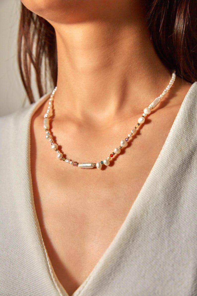 Op de afbeelding: Een ketting bestaande uit een streng witte zoetwaterparels afgewisseld met kleine, veelkleurige edelsteenkralen en piepkleine goudkleurige metalen tussenstukjes. De parels vari&euml;ren in vorm, waaronder ronde en langwerpige cilindrische vormen.