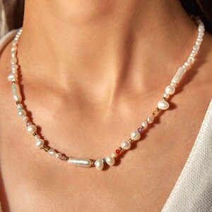 Op de afbeelding: Een ketting bestaande uit een streng witte zoetwaterparels in diverse vormen, waaronder ronde en langwerpige exemplaren. Tussen de parels bevinden zich kleine, goudkleurige metalen kralen en piepkleine, veelkleurige gefacetteerde accentkralen.