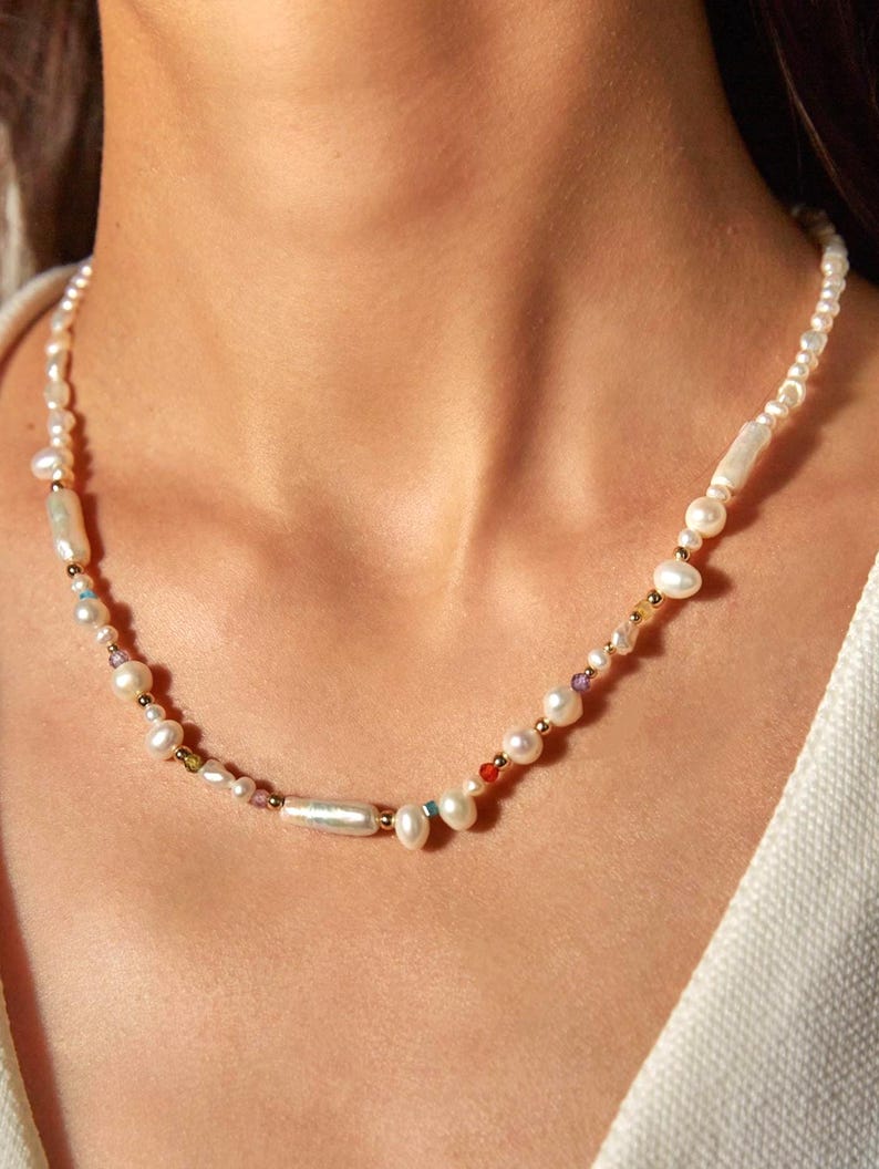 Op de afbeelding: Een ketting bestaande uit een streng witte zoetwaterparels in diverse vormen, waaronder ronde en langwerpige exemplaren. Tussen de parels bevinden zich kleine, goudkleurige metalen kralen en piepkleine, veelkleurige gefacetteerde accentkralen.
