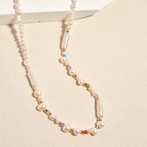 Op de afbeelding: Een ketting met een streng onregelmatige witte zoetwaterparels, afgewisseld met kleine goudkleurige kraaltjes en piepkleine, veelkleurige gefacetteerde edelsteenaccenten. Het ontwerp bevat langwerpige cilindrische parels en ronde parels.