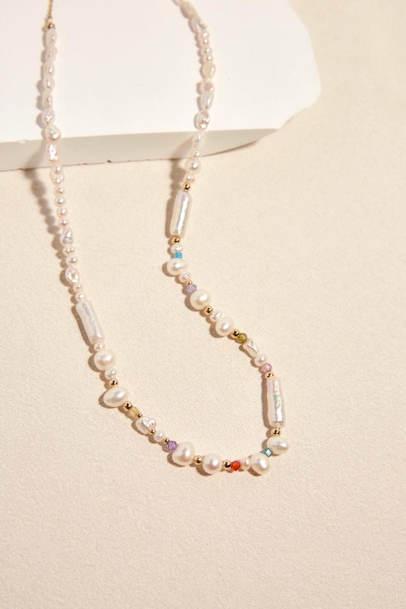 Op de afbeelding: Een ketting met een streng onregelmatige witte zoetwaterparels, afgewisseld met kleine goudkleurige kraaltjes en piepkleine, veelkleurige gefacetteerde edelsteenaccenten. Het ontwerp bevat langwerpige cilindrische parels en ronde parels.