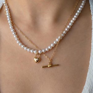 May include: A layered necklace featuring a strand of white pearls and a thin gold-tone chain. The piece includes a gold-tone heart-shaped padlock charm with a small pearl inlay and a gold-tone toggle clasp closure hanging from the chain.